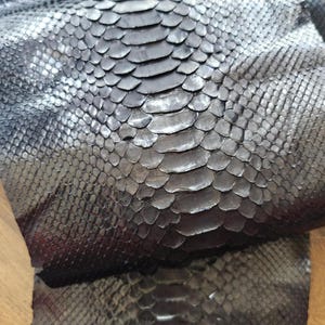 Könnte beinhalten: Nahaufnahme von dunkelgrauem Schlangenleder mit Schuppenmuster. Das Leder hat eine strukturierte Oberfläche und verschiedene Grautöne, die einen natürlichen, organischen Look erzeugen. Das Material scheint flexibel und für verschiedene Bastelprojekte geeignet zu sein.