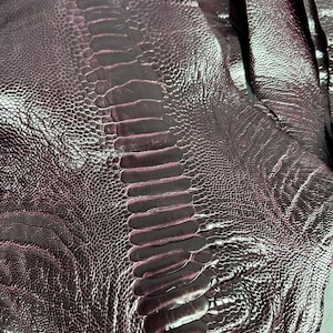 Op de afbeelding: Close-up van bordeauxkleurig struisvogelleer. Het leer heeft een getextureerd, korrelig oppervlak met een prominent centraal ruggengraatachtig patroon. De kleur varieert enigszins, met donkerdere tinten in de groeven en lichtere tinten op de verhoogde gebieden.