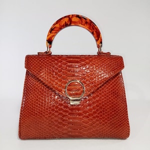 Può includere: Borsa a mano arancione con motivo a pelle di serpente. Ha un manico tartarugato e una chiusura circolare color oro. La chiusura a patta aggiunge un tocco classico a questo elegante accessorio.
