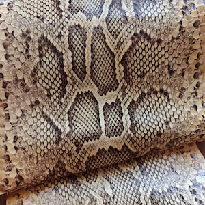 Puede incluir: Primer plano de cuero de piel de serpiente marrón y beige. El cuero tiene un patrón de escamas con una mezcla de tonos marrones claros y oscuros. La textura parece áspera y natural, con variaciones visibles en color y patrón.