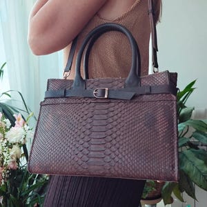 May include: A brown leather handbag with a snakeskin pattern. The bag has a top handle and a shoulder strap. A dark brown belt with a gold buckle wraps around the center. The bag is approximately 30 cm wide and 25 cm tall.