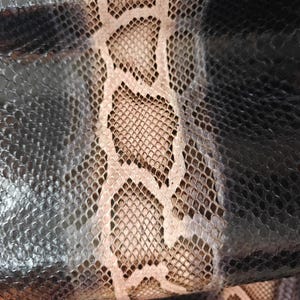 May include: Close-up of a snakeskin pattern. The image shows a detailed texture with a mix of dark grey, black, and tan scales. The pattern has a natural, organic appearance, with variations in colour and texture.
