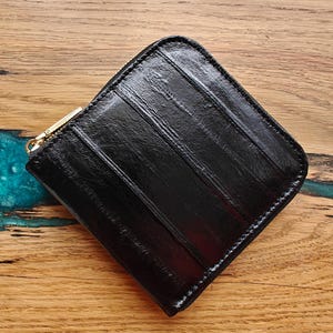 May include: A black, square-shaped wallet with a textured surface and a gold-colored zipper. The wallet is made of a shiny material and has vertical lines across the front. The wallet is on a wooden surface.