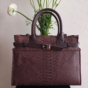 Könnte beinhalten: Eine braune Lederhandtasche mit Schlangenmuster. Die Tasche hat zwei Griffe, einen Gürtel mit goldener Schnalle und eine rechteckige Form. Weiße Blumen sind im Hintergrund.