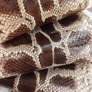 May include: Close-up of stacked, textured leather pieces. The leather displays a pattern of brown, tan, and cream scales, with a snake-like appearance. The scales vary in size and shape, creating a naturalistic texture.