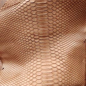 Op de afbeelding: Close-up van een bruin slangenvel met een geschubde textuur. De schubben variëren in grootte en vorm, waardoor een natuurlijk patroon ontstaat. De huid is gevouwen en toont de onderkant. De algehele kleur is een warme, aardse tint.