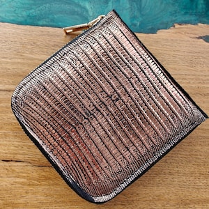 Pode incluir: Uma pequena carteira quadrada com acabamento em ouro rosa metálico e um padrão texturizado semelhante ao de um réptil. A carteira tem uma borda preta e um zíper dourado. O fundo é uma superfície de madeira.