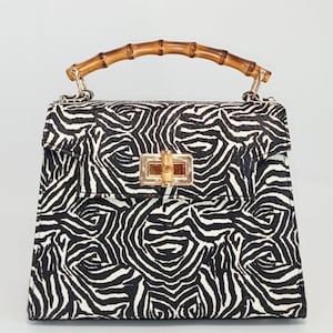 May include: A square handbag with a zebra print pattern in black and white. The bag features a bamboo handle and a gold-colored clasp. The bag is a stylish accessory.