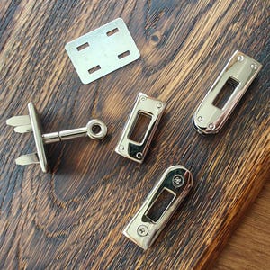 Puede incluir: Una variedad de componentes de ferretería metálicos plateados y brillantes, que incluyen cierres de cerradura rectangulares, una placa de montaje y un cierre con un pasador. Las piezas están dispuestas sobre una superficie de madera oscura y texturizada, lo que sugiere un proyecto de artesanía o bricolaje.