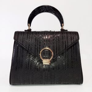 Puede incluir: Bolso de mano negro con un patrón texturizado similar al de un reptil. Cuenta con un asa curva y veteada y un cierre circular de color dorado. El bolso tiene una forma estructurada y un cierre de solapa.