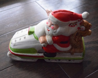 May include: A white, green, and red ceramic figurine of Santa Claus riding a snowmobile. Santa is wearing a red suit with white trim and a white beard. The snowmobile has a green seat and a white body.