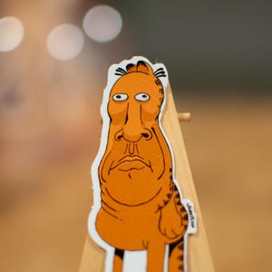 K&ouml;nnte beinhalten: Ein Aufkleber mit einer orangefarbenen Comicfigur mit einer langen Nase und gro&szlig;en Augen. Die Figur steht auf einer Holzoberfl&auml;che.