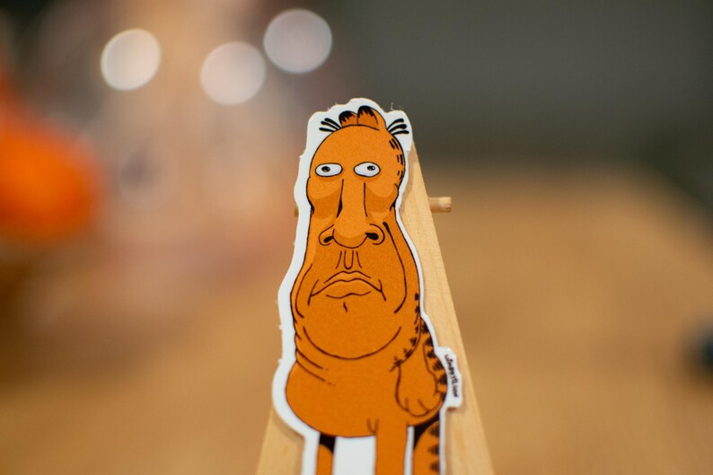 K&ouml;nnte beinhalten: Ein Aufkleber mit einer orangefarbenen Comicfigur mit einer langen Nase und gro&szlig;en Augen. Die Figur steht auf einer Holzoberfl&auml;che.