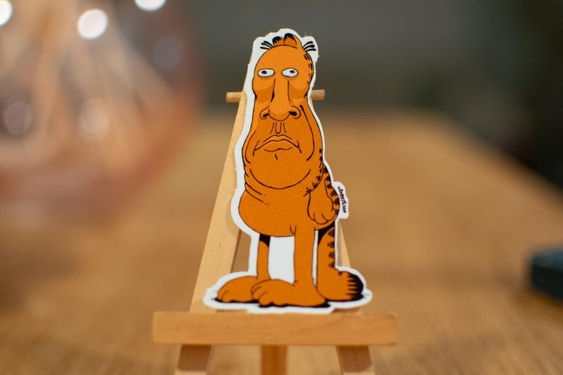 K&ouml;nnte beinhalten: Ein Aufkleber mit einer orangefarbenen Comic-Katze mit langem Gesicht und einem traurigen Ausdruck. Die Katze steht auf ihren Hinterbeinen und hat einen gestreiften Schwanz. Der Aufkleber befindet sich auf einem kleinen Holzstaffelei.