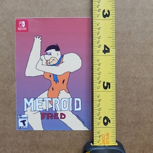 May include: A pink, purple, and blue Nintendo Switch game case featuring a cartoon illustration of Fred Flintstone in a pink and orange outfit. The text "METROID FRED" is printed on the case.