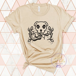 以下が含まれることがあります： ギターを弾いている犬の白黒のグラフィックが描かれたベージュのTシャツ。犬は真剣な表情をしていて、葉に囲まれています。Tシャツの下部には「good and bella BOUTIQUE」という文字が印刷されています。