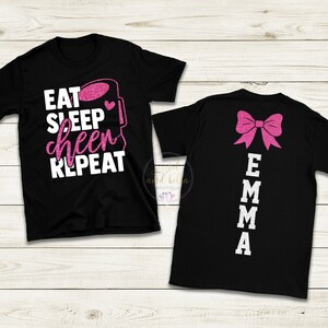 Könnte beinhalten: Zwei schwarze T-Shirts mit pinkfarbenen Glitzerdesigns. Das vordere Shirt hat den Text "Eat Sleep Cheer Repeat" mit einer pinkfarbenen Glitzer-Kaffeetasse. Das hintere Shirt hat eine pinkfarbene Glitzer-Schleife und den Text "Emma" vertikal auf dem Rücken.