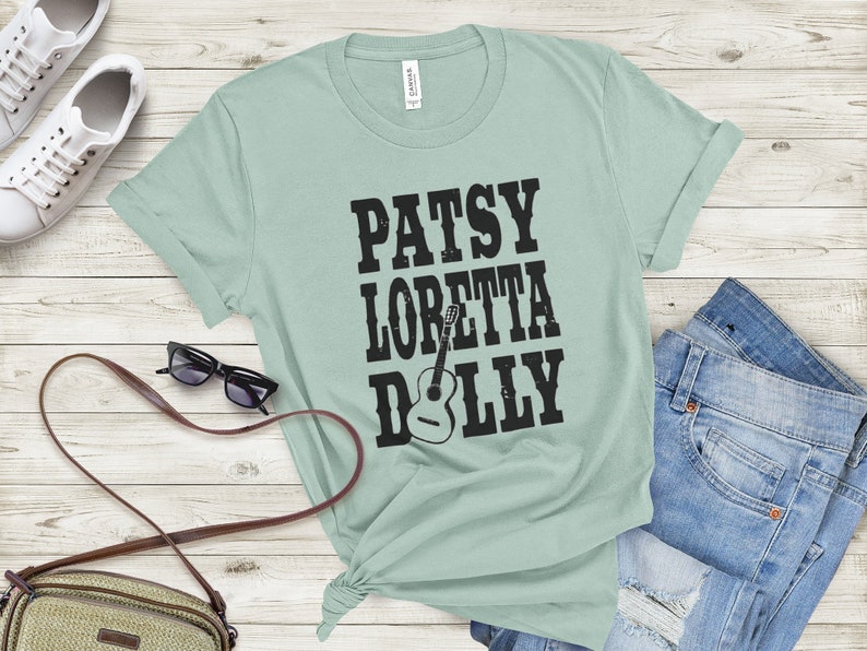 May include: A light green t-shirt with a black graphic print of the names "Patsy", "Loretta", and "Dolly" with a guitar silhouette.