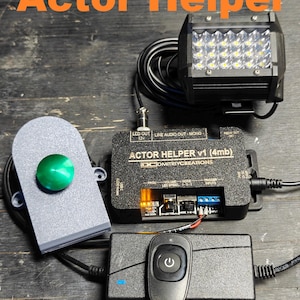 May include: An Actor Helper device with a black control box, a remote control, and a spotlight. The device has the text "Actor Helper" and "www.DmitriyCreations.com" printed on it. The spotlight has multiple LED lights.