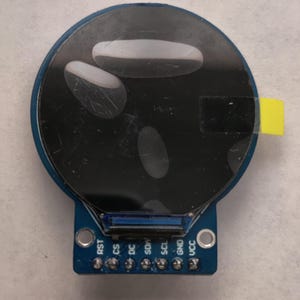 Könnte beinhalten: Ein kreisförmiges elektronisches Anzeigemodul mit einem dunklen Bildschirm, der drei blasse ovale Formen aufweist. Die blaue Leiterplattenbasis enthält sechs beschriftete Pins: RST, CS, DC, SDA, SCL, GND und VCC sowie zwei Montagelöcher und eine gelbe Klebelasche.