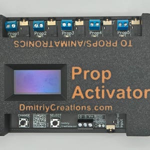 May include: A black Prop Activator with orange text and blue connectors. The device has an LCD screen and the text "DmitriyCreations.com". It is designed to connect to props and animatronics.