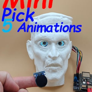 Puede incluir: Una cabeza esculpida blanca con ojos azules y el texto "Mini Pick 5 Animations" en rojo y azul. Un pequeño dispositivo circular negro está en un dedo, y una placa de circuito con cables es visible.