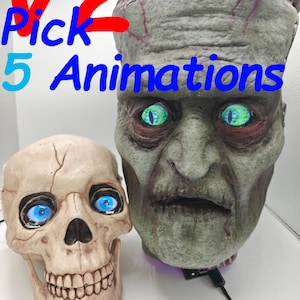 Op de afbeelding: Een Halloween-display met een schedel en een zombiehoofd. De schedel is gebroken wit met blauwe ogen en het zombiehoofd is groen met gloeiende groene ogen. De tekst "V2 Pick 5 Animations" staat in blauw en rood.