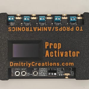 Könnte beinhalten: Ein schwarzer, rechteckiger elektronischer Prop Activator-Controller mit strukturierter Oberfläche. Er verfügt über einen zentralen Bildschirm, orangefarbene Beschriftungen, fünf blaue Klemmenblöcke, drei Steuertasten, einen QR-Code und einen Eingang für Bewegungssensoren.