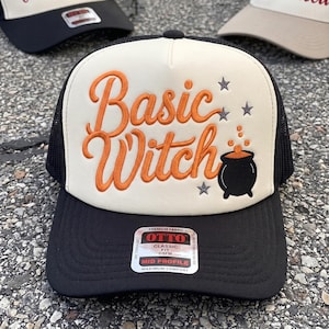 May include: A black and cream trucker hat featuring the embroidered orange text Basic Witch, a black cauldron graphic with orange bubbles, and three small grey stars. The front panel is cream, while the brim and mesh back are black.