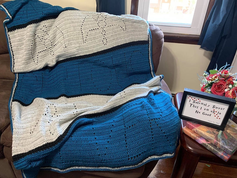 May include: A blue and white crocheted blanket with the word "Always" spelled out in the design. The blanket is folded and draped over a brown armchair. A framed quote reads "I solemnly swear that I am up to no good."