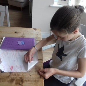 May include: A person wearing a white t-shirt with a black bird graphic is working at a wooden table. A purple folder with a sticker is on the table, along with a sheet of paper. The person is folding the corner of the paper.