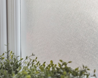 Könnte beinhalten: Eine Milchglasfolie mit einer strukturierten, weißen Oberfläche. Die Folie ist an einem Fenster mit einem weißen Rahmen angebracht. Im Vordergrund sind grüne Pflanzen zu sehen.