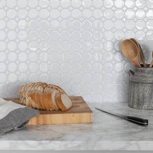 May include: A white octagon-shaped tile backsplash with a wooden chopping board and a loaf of sliced bread on top. A silver utensil holder with wooden spoons is on the right side of the image.