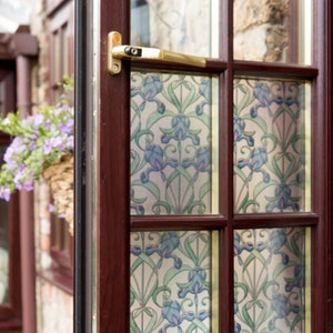 Könnte beinhalten: Teilweise geöffnetes Fenster mit dunklem Holzrahmen und einem dekorativen Blumenmuster auf dem Glas. Das Muster zeigt blaue und grüne irisartige Blumen. Ein goldfarbener Griff ist sichtbar. Ein hängender Korb mit lila Blumen ist zu sehen.