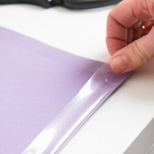 May include: A hand peeling a clear, adhesive strip from a sheet of light purple material. The material is on a white surface. The adhesive strip is partially lifted, revealing the surface beneath.
