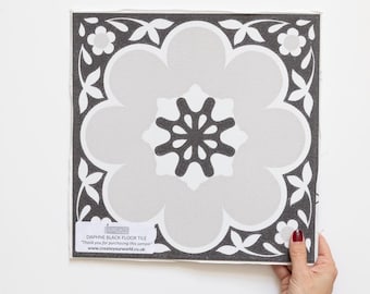 Könnte beinhalten: Quadratische Daphne Black Floor Tile Musterfliese mit floralem Design. Die Fliese hat eine große, hellgraue Blumenform in der Mitte, umgeben von einem dunkelgrauen Rand mit weißen floralen Akzenten. Der Text "Daphne Black Floor Tile" ist sichtbar.