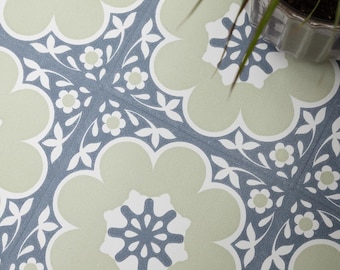 Könnte beinhalten: Nahaufnahme von gemusterten Bodenfliesen mit floralem Design. Die Fliesen weisen ein sich wiederholendes Muster aus großen, hellgrünen Blumenformen mit weißen und blauen Akzenten auf. Eine kleine Topfpflanze ist in der oberen rechten Ecke zu sehen.
