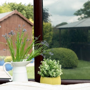 Puede incluir: Una jarra de cerámica blanca llena de lavanda y hierba verde se encuentra junto a una pequeña planta en maceta en una maceta amarilla tejida. Las plantas están sobre una superficie blanca frente a una ventana.