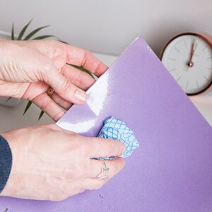 May include: A person's hands are holding a large, glossy, lavender-colored sheet of paper. A small, blue and white checkered cloth is used to wipe the surface. A copper-colored clock is in the background.