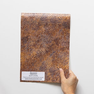 May include: A sample of "Rusty Stone" material with a textured, mottled appearance in shades of brown and orange. The sample is held up against a white background. A label at the bottom reads "Thank you for purchasing this sample" and a website address.