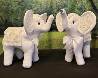 May include: Two white fabric elephant figurines with beaded embellishments and white satin bows. The elephants are facing each other.