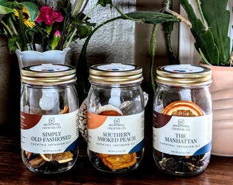 May include: Three glass jars with gold lids, each containing cocktail infusion kits. The jars have labels that read "Simply Old Fashioned," "Southern Smoked Peach," and "The Manhattan." The jars are filled with dried fruits and spices. The background includes flowers and plants.