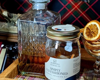 Puede incluir: Un decantador de vidrio transparente lleno de líquido ámbar se encuentra junto a un kit de infusión de cóctel Adornwell Cocktail Co. Simply Old Fashioned en un frasco de vidrio con tapa dorada, con rodajas de naranja seca y azúcar, 64 g.