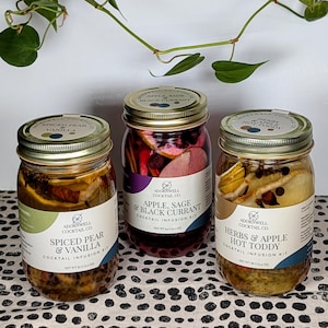 May include: Three glass jars with gold lids, each containing cocktail infusion kits. The jars are labeled "Spiced Pear & Vanilla", "Apple, Sage & Black Currant", and "Herbs & Apple Hot Toddy". The jars are filled with fruit and spices.