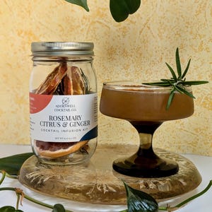 May include: A cocktail infusion kit with a glass jar containing dried citrus and rosemary, and a cocktail in a stemmed glass garnished with rosemary. The jar label reads "Rosemary Citrus & Ginger Cocktail Infusion Kit". The cocktail is on a decorative glass tray.