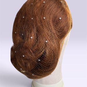Puede incluir: Un accesorio para el cabello que consiste en una red fina y transparente de color marrón sobre un moño de cabello castaño rojizo. La red está adornada con pequeños cristales transparentes espaciados que captan la luz con un brillo sutil.