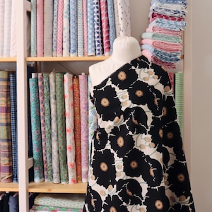 May include: A fabric bolt featuring a black floral pattern with tan centers and green accents is draped over a mannequin. Shelves behind the mannequin display a variety of colorful fabric rolls, showcasing a diverse selection of textiles.