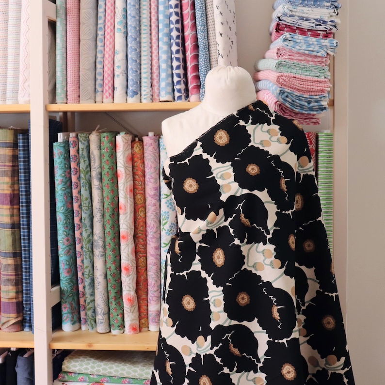 May include: A fabric bolt featuring a black floral pattern with tan centers and green accents is draped over a mannequin. Shelves behind the mannequin display a variety of colorful fabric rolls, showcasing a diverse selection of textiles.