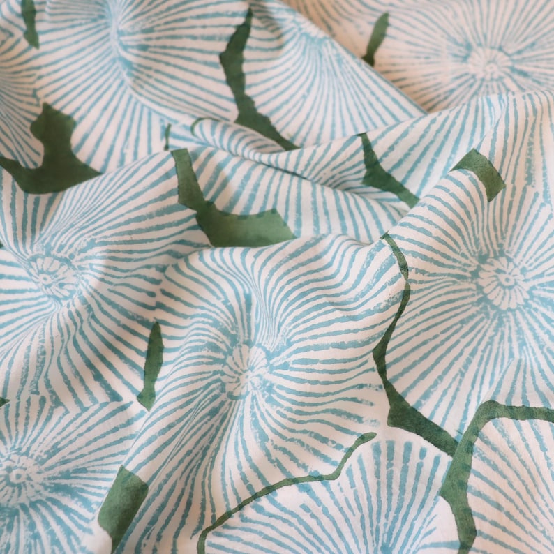 May include: A white fabric featuring a repeating pattern of light blue circular motifs with radiating lines. The design includes dark green organic shapes interspersed between the blue circular patterns, creating a textured and layered visual effect.