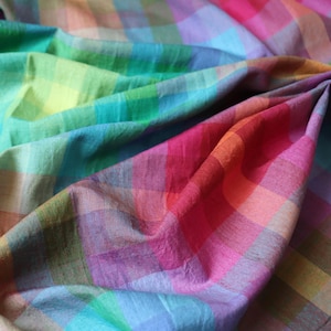 Puede incluir: Una tela ligera con un patrón de cuadros multicolor y vibrante. El tejido muestra una cuadrícula repetitiva de cuadrados en tonos rosa brillante, naranja, amarillo, verde, azul verdoso, azul y morado, con pliegues suaves que crean profundidad.